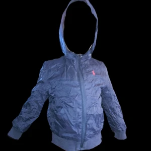 Svart vindjacka med huva - Säljer en mörkblå vindjacka med huva från Polo - Ralph Lauren. Jackan har en dragkedja framtill och är långärmad. Perfekt för blåsiga dagar och har en cool, sportig look. Passar bra till både vardag och lättare utomhusaktiviteter. Storlek 5 år