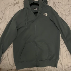 Grå hoodie från The North Face - Säljer en snygg och bekväm grå hoodie från The North Face. Den har en dragkedja framtill och en justerbar huva med snören. Perfekt för kyliga dagar och har en klassisk passform. Den är i bra skick.