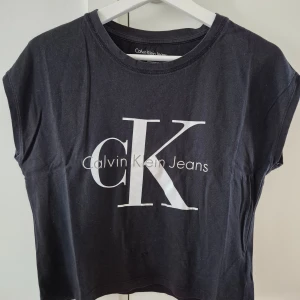 Tshirt CK - Svart T-shirt med vit och grå tryck från Calvin Klein. Lös modell. Storlek S. 100% bomull. Det är 3 st små stygn vid magen pga små hål. 