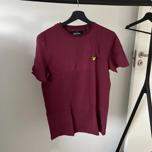 Vinröd t-shirt från Lyle & Scott - Säljer en snygg vinröd t-shirt från Lyle & Scott. Den har en klassisk passform och är kortärmad, perfekt för både vardag och fest. T-shirten är i bomull och har en broderad gul logga på bröstet. Skön och stilren, ett måste i garderoben!