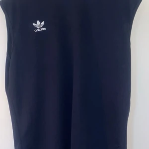 Ärmlös tröja från Adidas - Denna ärmlösa tröja från Adidas är tillverkad i ett tunt och lätt tyg, vilket gör den idealisk för fysisk aktivitet eller en avslappnad look. Den har en elegant mörk marinblå färg, vilket gör den till ett mångsidigt plagg som är lätt att matcha. 
