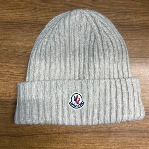 Moncler Mössa Beige - Snygg grå ribbad mössa från Moncler med broderad logotyp framtill. Perfekt för kyliga dagar och ger en stilren look. Tillverkad i mjukt material för extra komfort. Pris sänkes vid snabbt köp