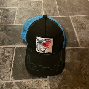 Svart och blå keps med hajmotiv - Cool keps med svart front och blått mesh-tyg på sidorna. Framtill finns ett broderat hajmotiv som ger en unik touch. Perfekt för en avslappnad stil.