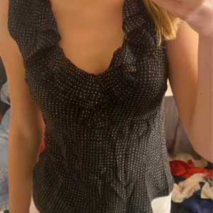 Svart prickig blus med volangdetaljer - Snygg svart blus med vita prickar och volangdetaljer vid halsringningen. Perfekt för sommaren!!!