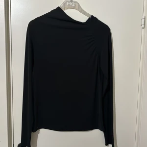 Svart långärmad topp från Visual Clothing Project - Snygg svart långärmad topp från Visual Clothing Project. Den har en stilren design med en lätt drapering vid axeln som ger en unik touch. Perfekt för en minimalistisk look.