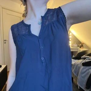 Mörkblå ärmlös blus med spetsdetaljer - Snygg mörkblå blus med spetsdetaljer på axlarna och knappar framtill. Perfekt för en stilren look. Blusen är ärmlös och har en avslappnad passform.❤️