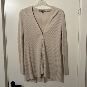 Beige ribbad kofta från Esprit - Snygg beige kofta från Esprit med ribbad design och v-ringning. Den har långa ärmar och knappar framtill, perfekt för lager-på-lager-stil. Passar både till vardags och mer uppklädda tillfällen.