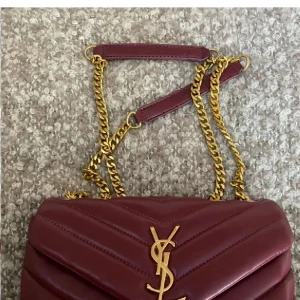 Vinröd axelväska från YSL - Snygg vinröd axelväska från YSL i skinn med quiltad design och guldfärgade kedjedetaljer. Väskan har en elegant YSL-logga på framsidan och en praktisk dragkedja inuti. Perfekt för att ge din outfit en lyxig touch. Dock ingen aning om den är äkta. Finns inget äkthetsbevis. Lägger ut igen då man får gratis frakt. 