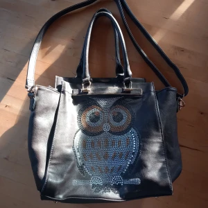 Väska med ugglemotiv - Snygg svart axelväska i syntetmaterial med ett ugglemotiv på framsidan. Väskan har både handtag och en justerbar axelrem för flexibel användning. Inga skador eller defekter. 