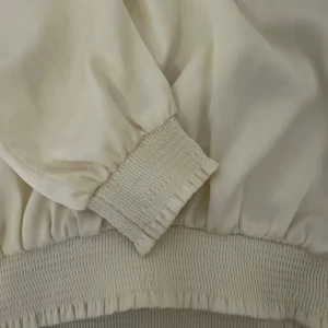 Vit blus från Gina Tricot - Säljer en stilren vit blus från Gina Tricot med elastiska detaljer vid hals, ärmslut och nederkant. Blusen har långa ärmar och en luftig passform, perfekt för en chic look. Passar till både vardag och speciella tillfällen.