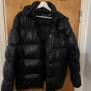 Ralph Lauren jacka - Säljer en snygg svart dunjacka från Ralph Lauren. Jackan har en klassisk design med dragkedja och en liten broderad logotyp på bröstet. Perfekt för kyliga dagar. 