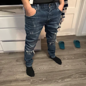Blå slitna jeans - Säljer ett par blå jeans med slitningar och en klassisk femficksdesign. Jeansen har en dragkedja och knapp framtill. Perfekta för en avslappnad stil.