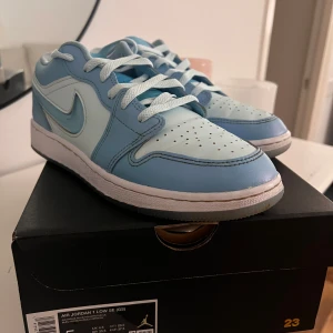 Nike Air Jordan 1 Low i ljusblått - Snygga Nike Air Jordan 1 Low sneakers i en fräsch ljusblå färg med mörkblå detaljer. Skorna har klassisk snörning och perforeringar på tån för extra ventilation. Perfekta för en stilren look. Skickas i original förpackning. 