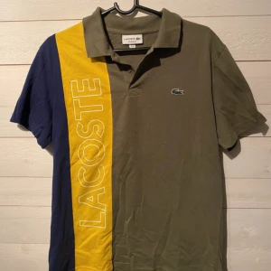 Grön och gul kortärmad pikétröja från Lacoste - Snygg grön pikétröja från Lacoste med korta ärmar och en iögonfallande gul och blå panel med stor logotyp. Klassisk krage och broderad krokodil på bröstet. Perfekt för en sportig look!