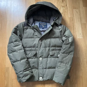 Grön dunjacka från Woolrich - Säljer en grön dunjacka från Woolrich med huva och knappar. Jackan har en quiltad design och är perfekt för kyliga dagar. Den är i grymt skick! 