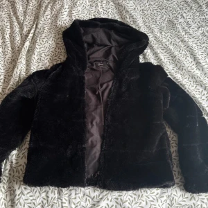 Svart pälsjacka med huva - Mysig svart pälsjacka med huva, perfekt för kyliga dagar. Jackan har en mjuk och fluffig yta som ger en lyxig känsla. Den är långärmad och har en enkel design utan synliga detaljer som dragkedjor eller knappar.