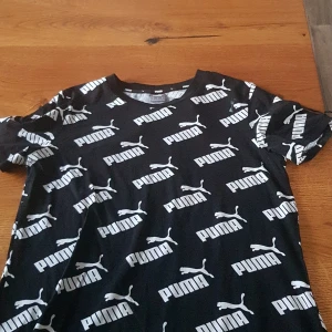 Svart t-shirt/klänning från Puma - Snygg svart t-shirt från Puma med ett iögonfallande vitt logotypmönster över hela plagget. Perfekt för en sportig och avslappnad stil.