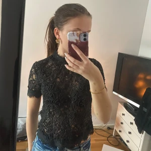 Svart spetströja med dragkedja - Säljer en snygg svart spetstopp med korta ärmar och dragkedja baktill. Perfekt för en festlig kväll eller som ett stiligt lager över en enkel topp. Toppen har en elegant och luftig design som ger en chic look.