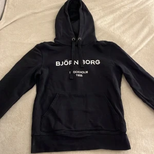 Svart hoodie från Björn Borg - Säljer en svart hoodie från Björn Borg. Den har en klassisk känguruficka och justerbar huva med dragsko. Perfekt för en avslappnad stil eller mysiga dagar hemma. 🖤