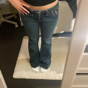 Bootcut lågmidjade True Religion i nyskick - Snygga mörkblå bootcut jeans från True Religion med ikoniska bakfickor och kontrastsömmar. Jeansen har låg midja och en klassisk femficksdesign med knappar och dragkedja framtill.