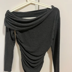 Mörkgrå offshoulder topp - Snygg mörkgrå offshoulder topp med draperad design. Perfekt för en stilren look med långa ärmar och mjukt material.