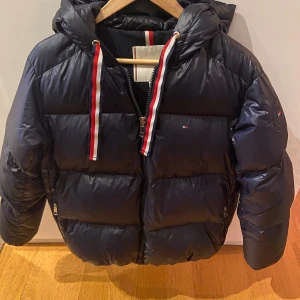 Marinblå dunjacka med huva - Snygg marinblå dunjacka från Tommy Hilfiger med huva och dragkedja. Jackan har röda och vita detaljer på snörena och en liten logotyp på ärmen. Perfekt för kyliga dagar.