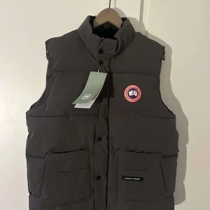 Grå dunväst från Canada Goose - Säljer en stilren grå dunväst från Canada Goose med broderad logotyp på bröstet. Västen har en hög krage och knappar framtill. Perfekt för kyliga dagar.  Kom med prisförslag