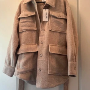 Beige jacka med bälte från NA-KD - Säljer en stilren beige jacka från NA-KD i storlek EU 40. Jackan har ett bälte i midjan och stora fickor framtill. Perfekt för en trendig look!