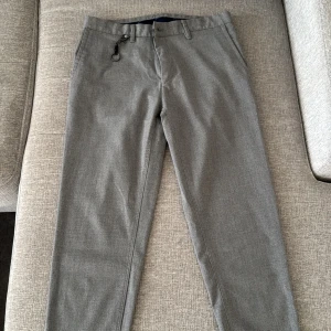 Grå kostymbyxor med knappdetaljer - Snygga grå kostymbyxor med stilrena knappdetaljer och praktiska fickor. Byxorna har en klassisk passform och är perfekta för en elegant look. De är utrustade med bälteshällor och en diskret dragkedja framtill.