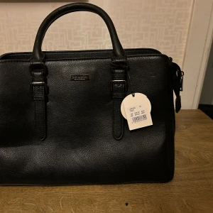 Svart handväska från Don Donna - Snygg svart handväska från Don Donna i skinn med stilrena detaljer och dubbla handtag. Perfekt för att bära dina dagliga nödvändigheter med stil. Väskan har en praktisk dragkedja för säker förvaring.