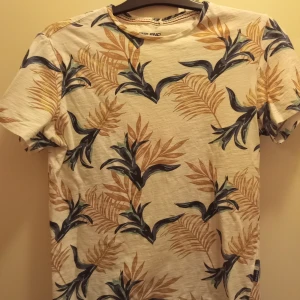 Vit t-shirt med bladmönster från Blend - Säljer en vit t-shirt från Blend med ett tropiskt bladmönster. T-shirten har korta ärmar och är perfekt för en avslappnad stil.