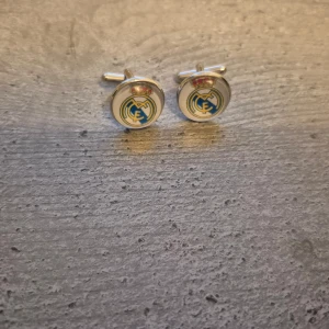 Manschettknappar med Real Madrid-logga - Snygga manschettknappar med Real Madrid-logga i blått och gult på en silverfärgad bas. Perfekta för fotbollsfans som vill visa sitt stöd med stil.