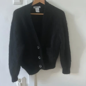 Svart kofta från Hope - Mysig svart kofta från Hope med knappar framtill. Den har en fluffig textur och långa ärmar, perfekt för kyligare dagar. En stilren och enkel design som passar till det mesta.