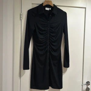 Svart, tight klänning - Nyskick. Ribbat material, stretchig. Snygga rynkor fram och med knappar och krage. Använd med tex en tröja över för en vardagligare look eller styla till fest. 