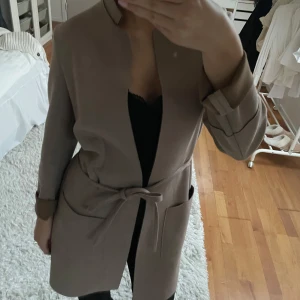 Beige kappa från H&M - Elegant beige kappa från H&M med knytband i midjan och långa ärmar. Perfekt för höst och vår, med en stilren design utan krage.