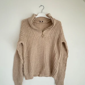 Beige stickad tröja från b.young - Mysig beige stickad tröja från b.young med en hög krage och dragkedja. Perfekt för kyliga dagar med sin ribbade struktur och långa ärmar. En stilren och bekväm tröja för alla tillfällen.