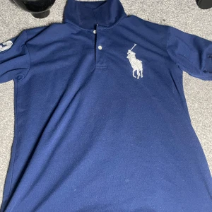 Mörkblå pikétröja från Ralph Lauren - Säljer en snygg mörkblå pikétröja från Ralph Lauren med klassisk krage och knappar. Tröjan har en stor vit logotyp på bröstet och en siffra på ärmen. Perfekt för en stilren look!