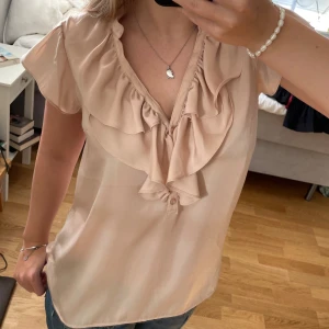 Beige blus med volangdetaljer - Säljer en superfin beige blus med volangdetaljer framtill. Den har korta ärmar och en v-ringad design som ger en elegant touch. Perfekt för både vardag och fest!😍😍