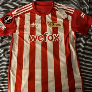 Union Berlin fotbolls tröja  - Säljer en äkta och oanvänd Union Berlin hemma tröja från säsongen 2022/2023 med tag! Tröjan har en pyte liten smuts fläk längst nere till vänster på baksidan av tröjan som säkert går bort i tvätten! Storlek:M ny pris:1000 kr  pris kan diskuteras! 