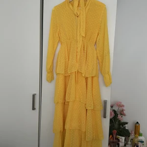 Gul långklänning med volanger - Säljer en superfin gul långklänning med volanger och prickigt mönster. Klänningen har långa ärmar och knappar framtill, samt en söt knytning vid halsen. Perfekt för sommarens fester eller en solig dag ute! 🌼