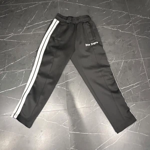  Palm Angels byxor - Snygga svarta mjukisbyxor från Palm Angels med vita ränder längs sidorna. Byxorna har en elastisk midja och en avslappnad passform, perfekt för en casual look.