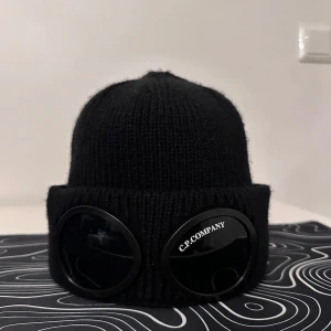 Svart mössa med glasögondetalj från C.P. Company - Snygg svart mössa från C.P. Company med unika glasögondetaljer framtill. Tillverkad i 100% bomull för en bekväm passform. Perfekt för att ge din outfit en cool touch.