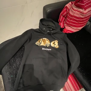 Svart hoodie från Palm Angels - Säljer en svart hoodie från Palm Angels med ett unikt nallebjörnsmotiv på framsidan. Tröjan har en klassisk känguruficka och justerbar huva. Perfekt för en avslappnad stil.