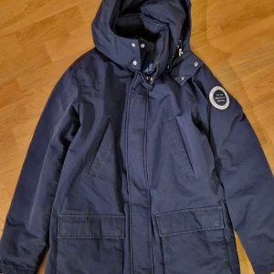 GANT Mörkblå parkajacka med huva - Snygg fodrad GANT mörkblå parkajacka med huva och praktiska fickor. Jackan har en stilren design med knappar och dragkedja framtill. Perfekt för kyligare dagar. 100% äkta eftersom den köpts i GANT butik. Name tag på fodret har överstruket namn. Djur- och rökfritt hem. 