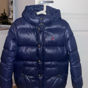 Blå dunjacka från Ralph Lauren - Snygg blå dunjacka från Ralph Lauren med huva och knappar framtill. Jackan har en klassisk design med broderad logotyp på bröstet. Perfekt för kyliga dagar.
