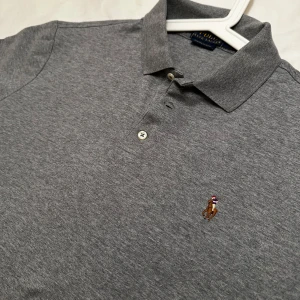 Grå pikétröja från Ralph Lauren - Säljer en klassisk grå pikétröja från Ralph Lauren med den ikoniska broderade logotypen på bröstet. Tröjan har en krage med två knappar och korta ärmar. Perfekt för en stilren och avslappnad look. Slim fit Large så sitter som en M. Helt ny endast testad.