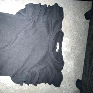 Svart topp med volangdetaljer från Zara - Säljer en stilren svart topp från Zara med volangdetaljer på axlarna. Perfekt för att ge en extra touch till din outfit. Toppen är i en bekväm passform och passar till många olika tillfällen.