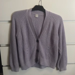 Lila stickad kofta från Lindex - Säljer en mysig lila stickad kofta från Lindex. Koftan har en v-ringad design med knappar framtill och ribbade muddar. Perfekt för kyliga dagar! Den har dock ett litet håll där bak, som nu ser på bilden.🥰