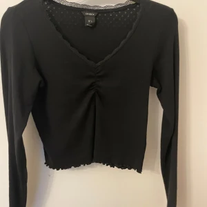 Svart långärmad topp från Lindex - Snygg svart långärmad topp från Lindex med v-ringning och spetsdetaljer. Toppen är croppad och har en ribbad kant vid ärmslut och nederkant. Perfekt för en stilren look.