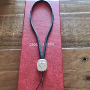 Georg Jensen mobil/väsk charm med hjärtdetalj - Elegant charm från Georg Jensen med en silverfärgad berlock i form av  ett hjärta. Smycket är fäst på ett svart läderband, vilket ger en stilren och tidlös look. Perfekt för att ge en touch av klass till vilken outfit som helst. Hjärtat i Sterling silver 925. Aldrig använt men behöver puts.